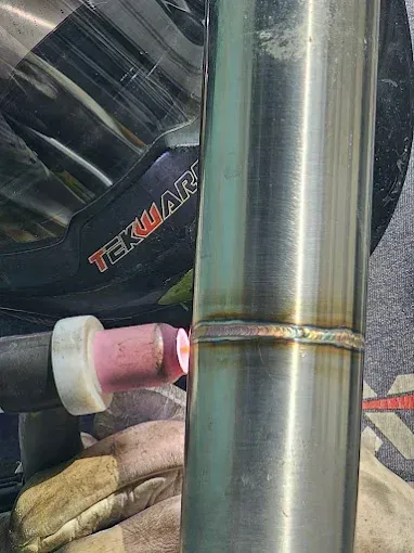 A TIG welder uses a torch to perform a precision weld on a vertical stainless steel pipe while wearing protective gear.