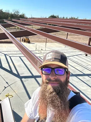A person with a long beard and glasses wearing a cap smiles while taking a selfie on a construction site with steel beams.