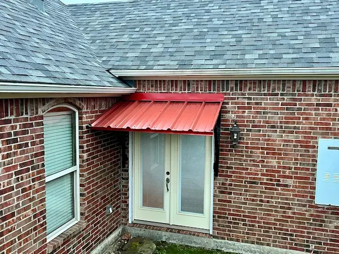 A red metal awning hangs above beige double doors on a brick house with a gray shingled roof.