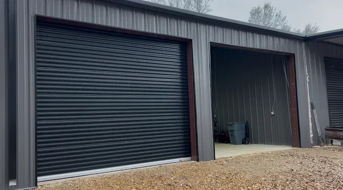 A grey metal storage building with a closed black roll-up door on the left and an open bay on the right.