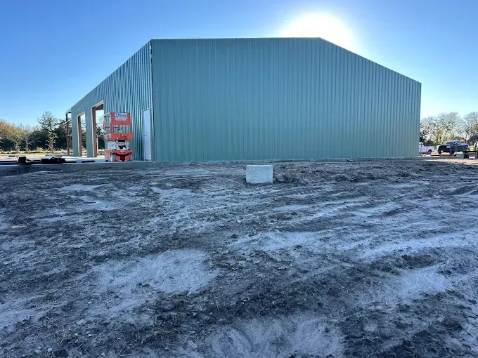 A large, light green metal building under construction on a dirt lot with a red scissor lift near the open entrance.