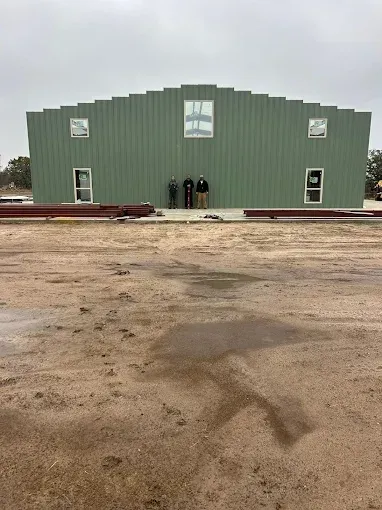 Three people stand in front of a green metal-sided building with windows, situated on a dirt lot under an overcast sky.