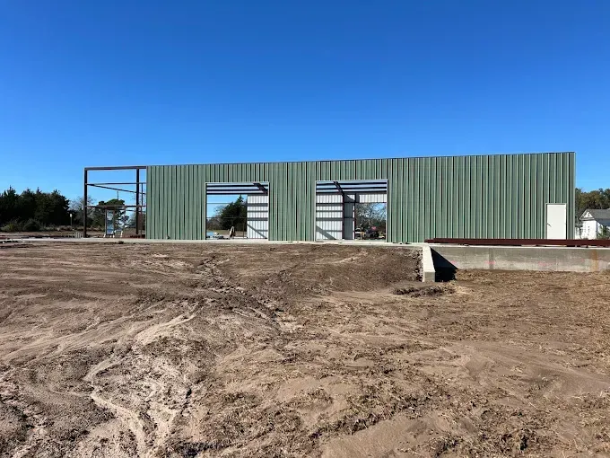 A wide shot of a green metal warehouse under construction on a dirt lot against a clear blue sky.