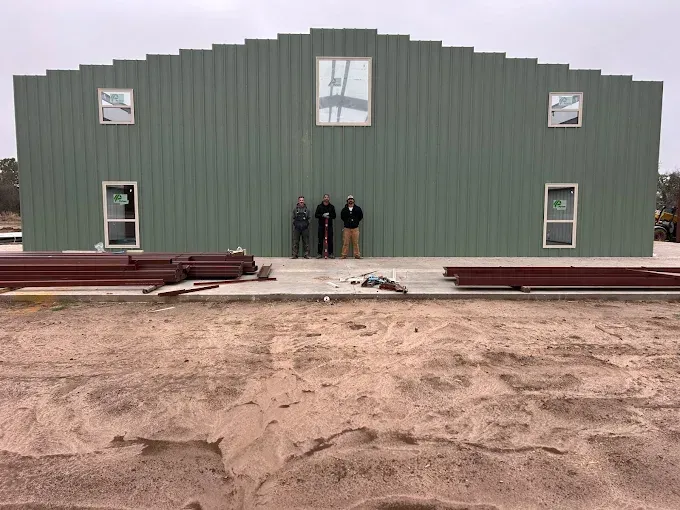Three people stand before a large, green metal-sided building under construction with windows and piles of steel beams.