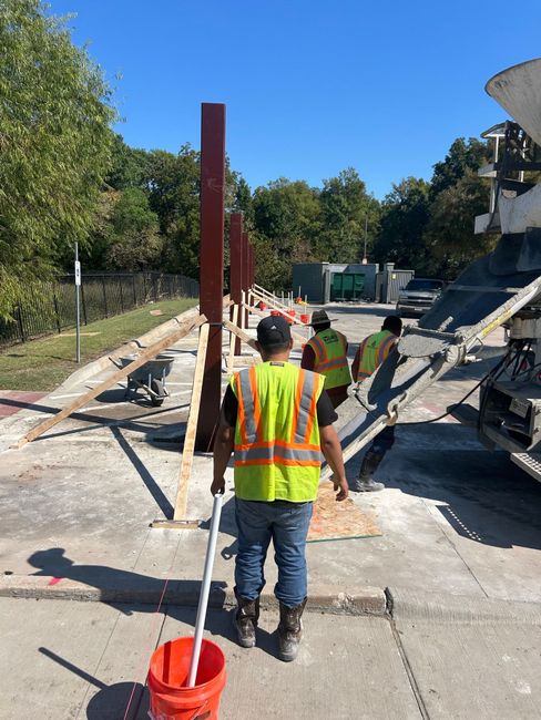 Construction workers pour concrete around vertical steel posts at an outdoor job site.