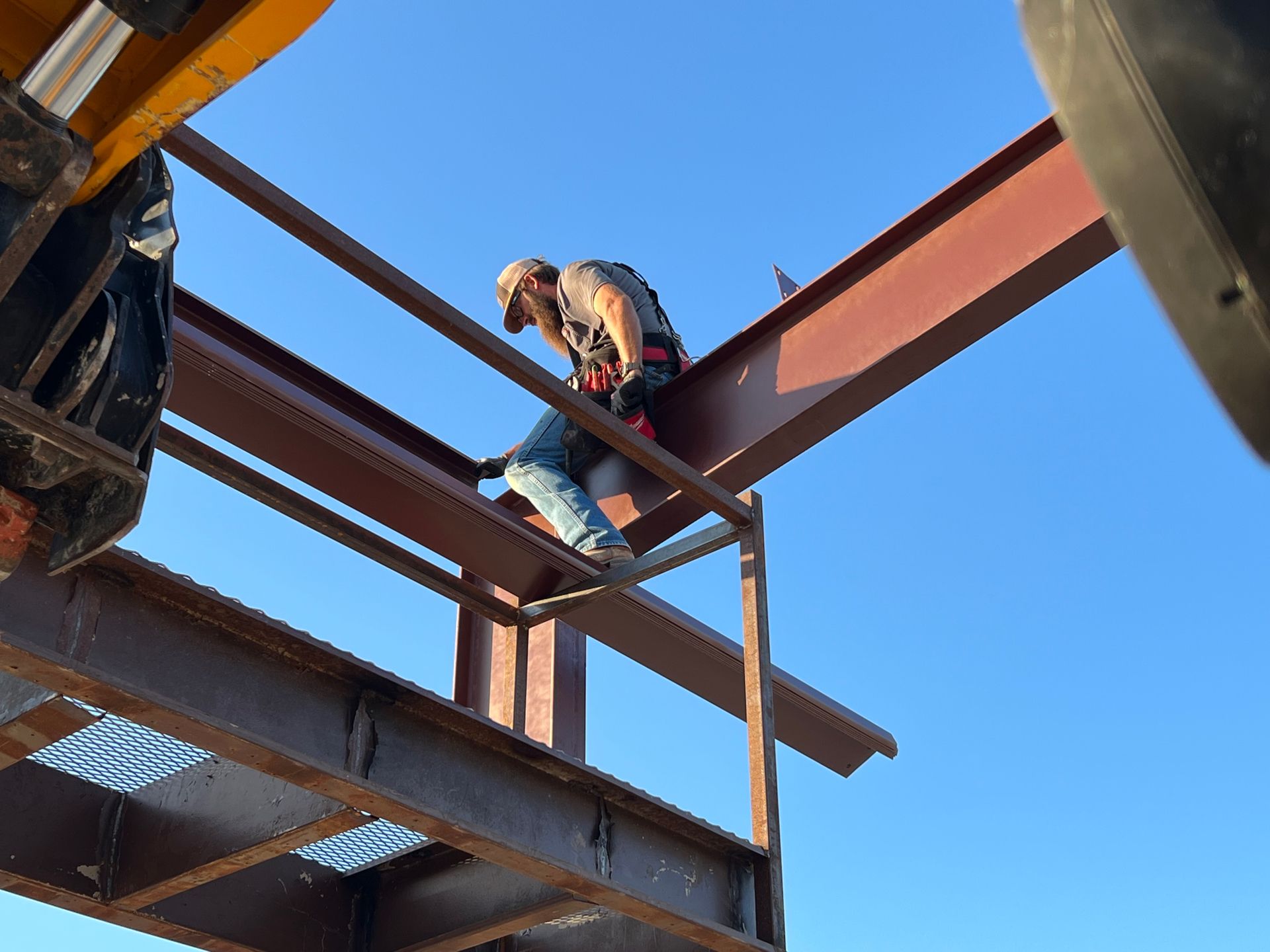 A construction worker in a safety harness sits on a brown steel beam against a clear blue sky.