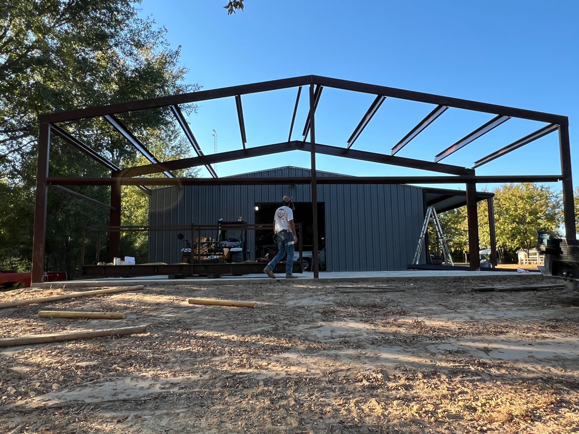 A person walks in front of a metal building frame under construction in an outdoor, wooded setting.