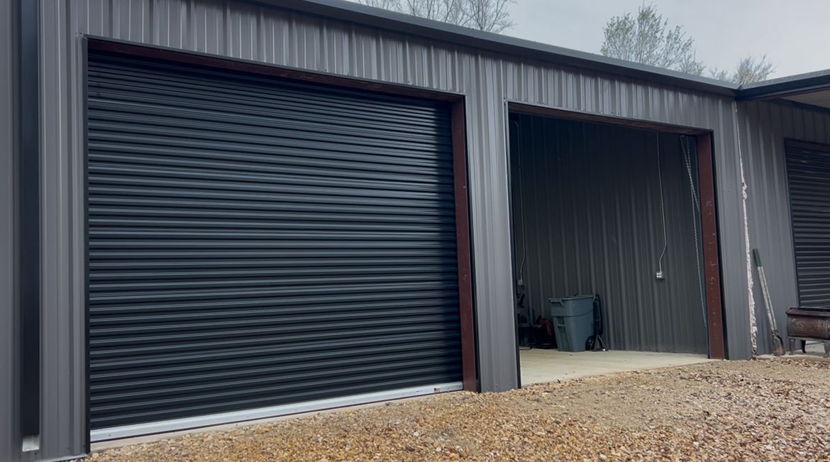 A gray metal garage building with one closed black roll-up door and one open bay on a gravel lot.