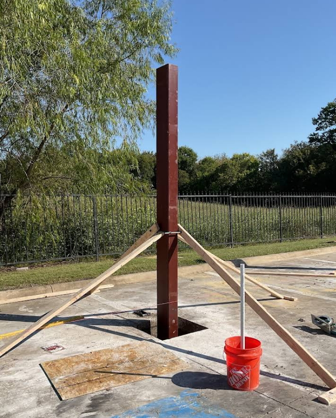 A steel support column held upright by wooden braces over an open square hole in a concrete surface.