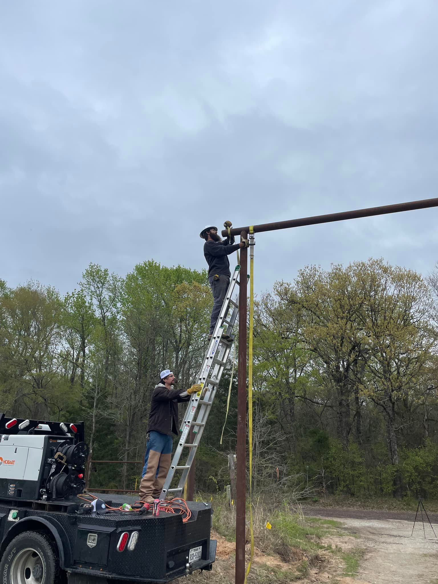 Two workers in work attire use an extension ladder to reach a horizontal pole, with one worker secured to the pole.