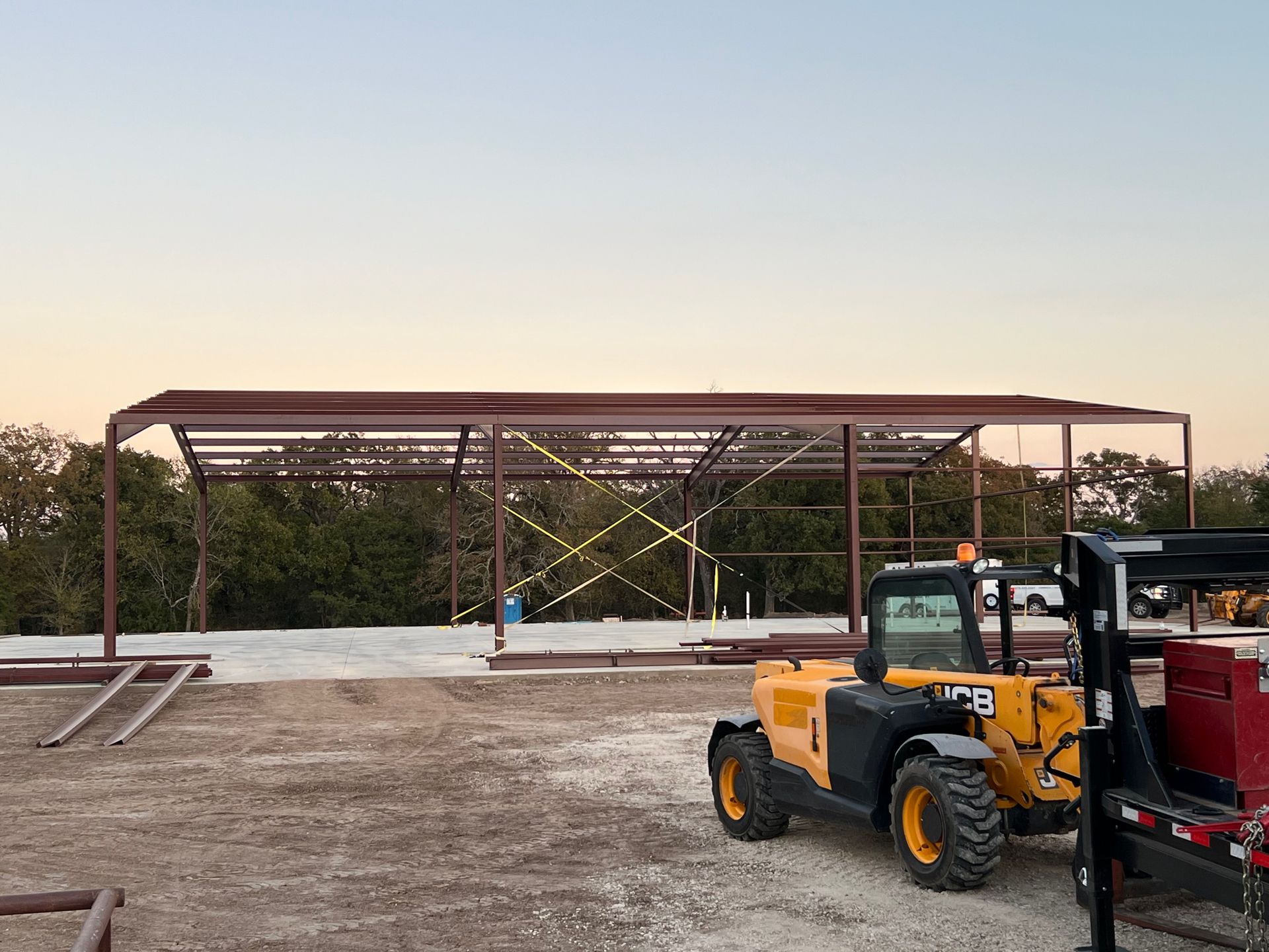 A metal frame for a building under construction, with a yellow telehandler parked on gravel in the foreground at dusk.