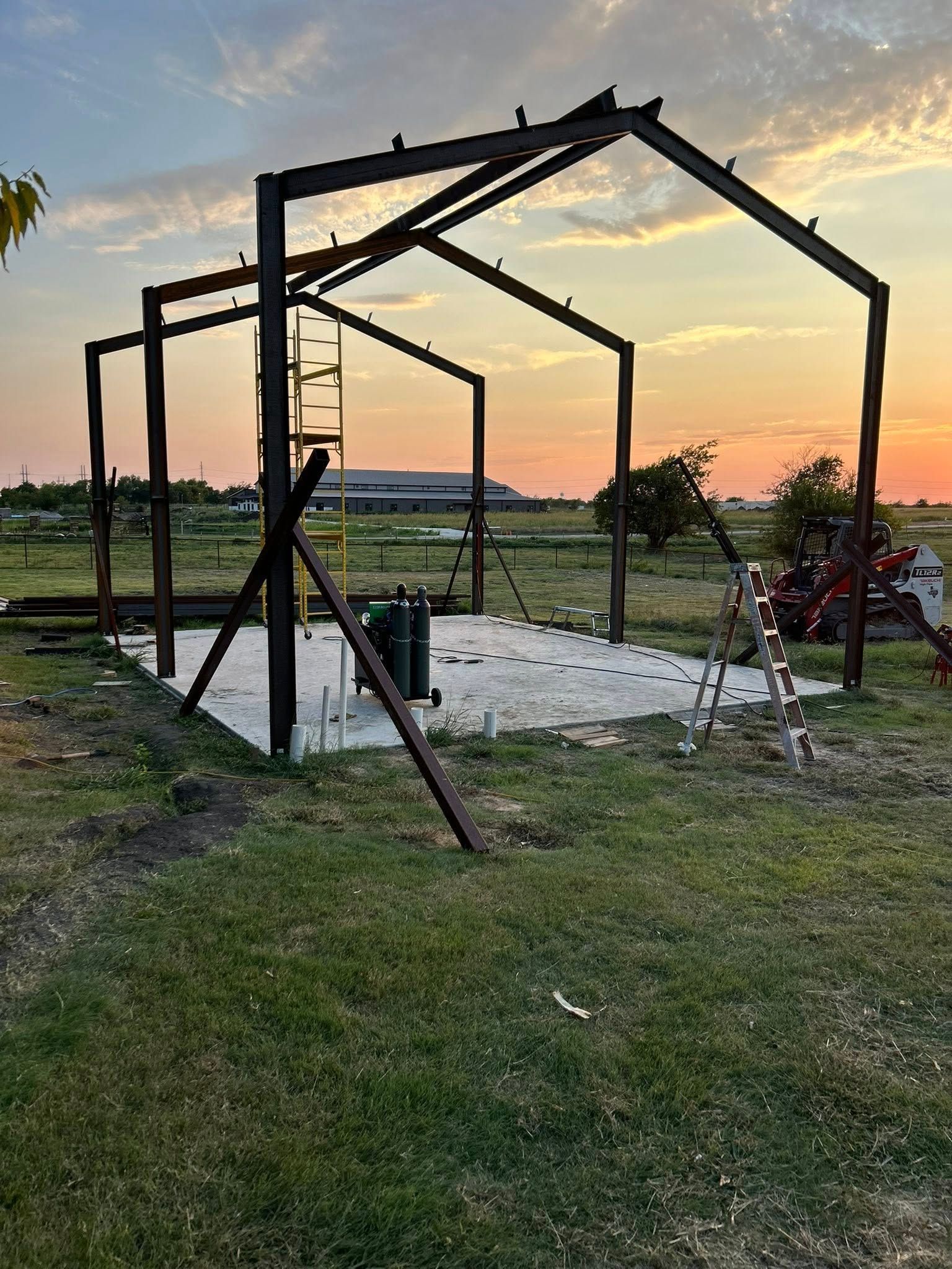 A steel frame for a building under construction on a concrete slab in an open field at sunset.