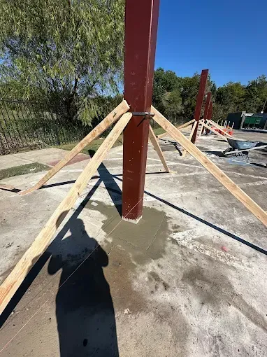 A vertical red steel post set in a fresh concrete base, held upright by diagonal wooden supports on a paved lot.