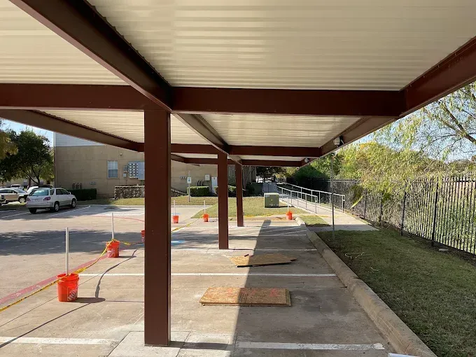 Outdoor covered parking spaces with metal structure and roof, orange buckets marking spots, and concrete ground.