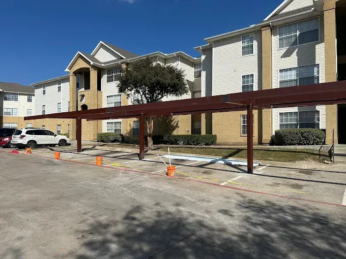 An apartment building exterior with a tan-colored metal carport structure in the foreground and a parked white SUV.
