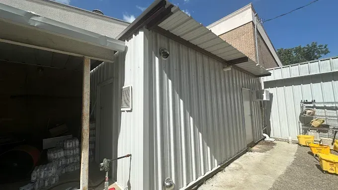 A white corrugated metal storage building exterior under a clear blue sky, with a covered entryway and outdoor buckets.