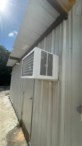A white window air conditioning unit mounted on the exterior corrugated metal wall of a building under a sunny sky.