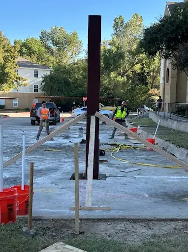 Two workers in high-visibility vests stabilize a tall, dark metal post with wooden braces on a parking lot concrete pad.