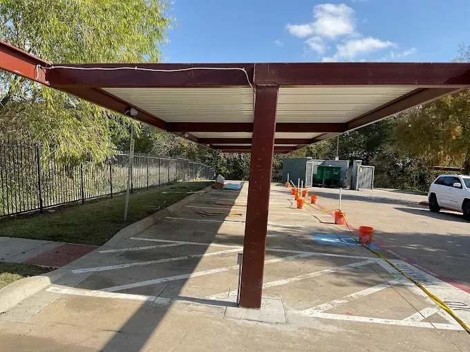 A brown metal parking carport structure with a white roof canopy over an asphalt lot with white painted stall lines.