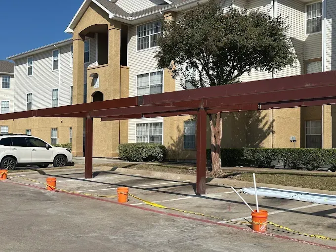 A partially constructed metal carport frame stands in an apartment complex parking lot near orange buckets and yellow tape.