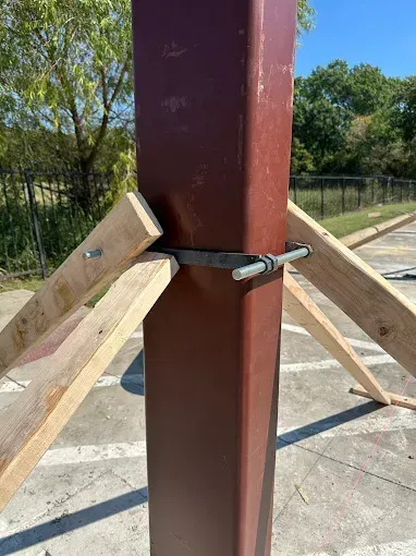 A red metal post held upright by wooden bracing supports attached with a metal band clamp on an outdoor concrete surface.