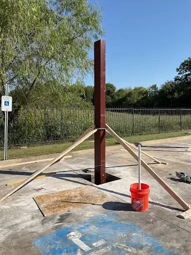 A tall, brown metal post stands in an outdoor parking lot, supported by wooden braces, with a red bucket nearby.