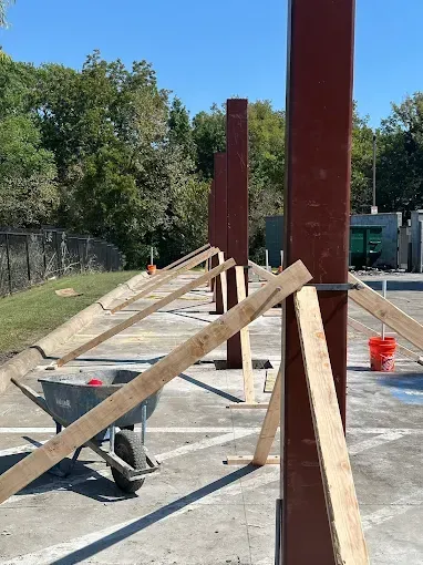 A row of brown steel construction beams standing upright on a paved surface, supported by wooden diagonal bracing.