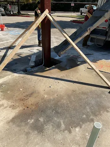 Workers use a wooden frame to brace a metal pole while concrete is poured into a hole in a parking lot.