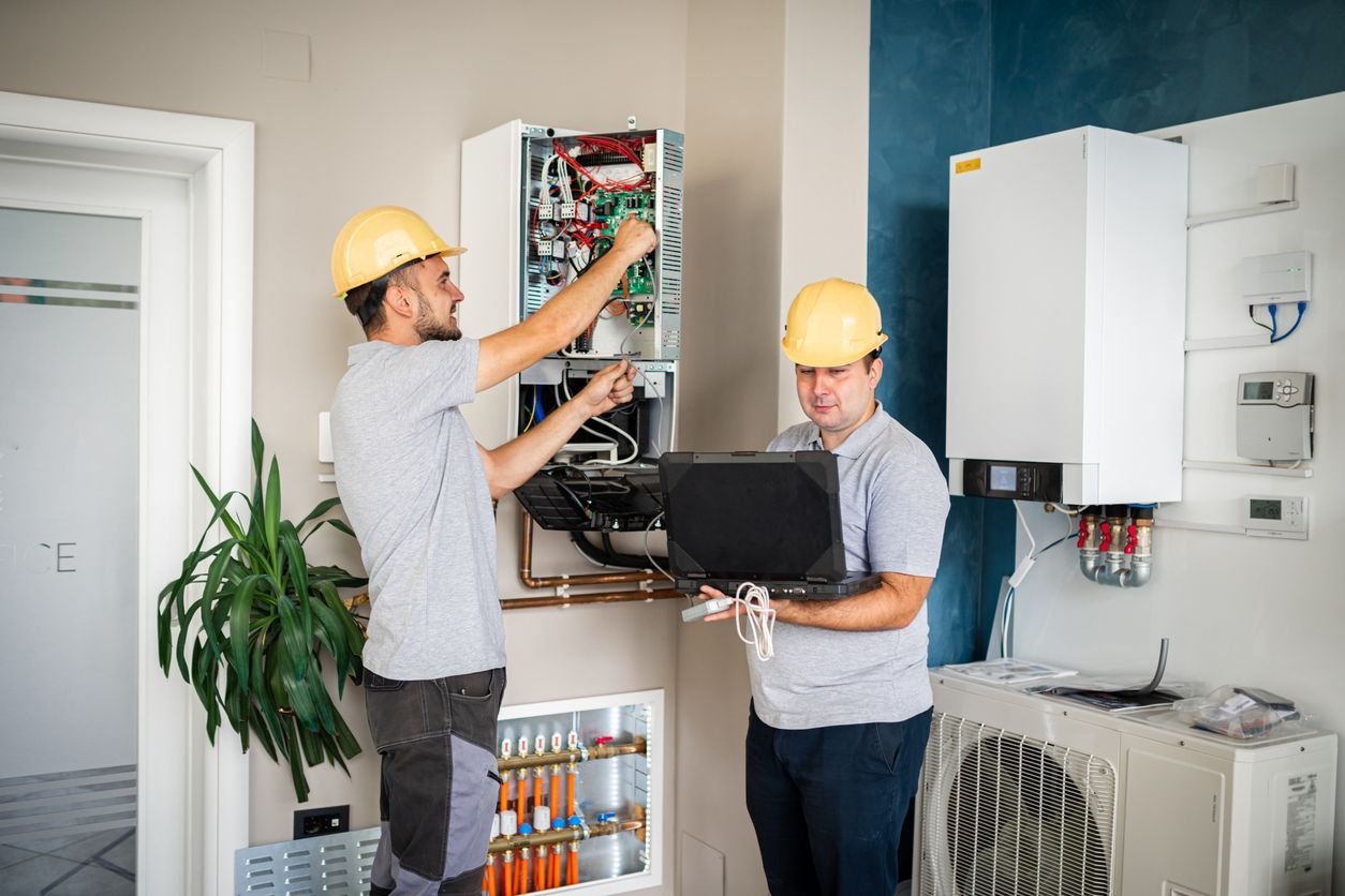Two technicians in hard hats inspect electrical panels in a utility room, one holding a laptop.
