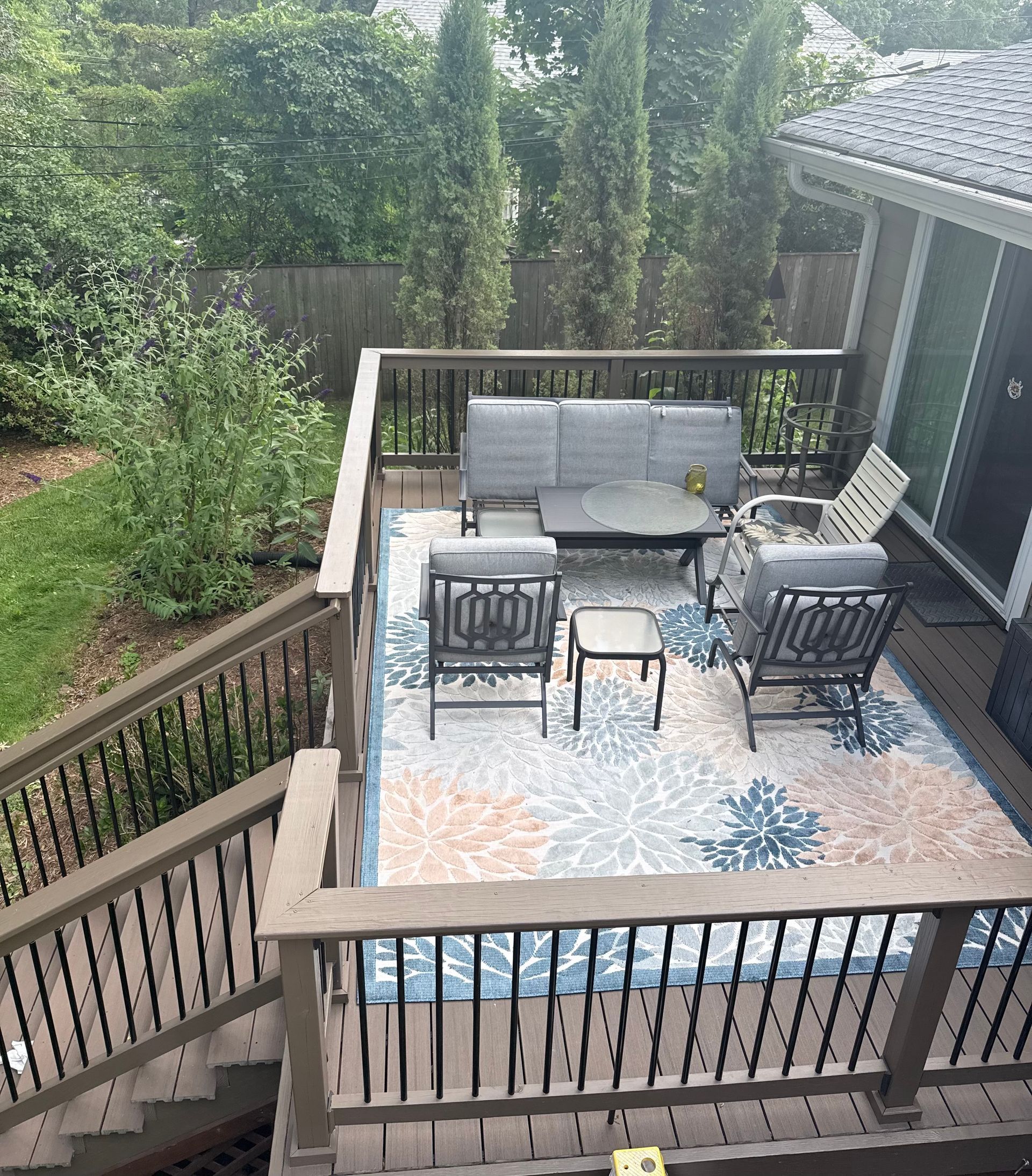Outdoor deck with gray seating, patterned rug, and stairs overlooking a wooded backyard