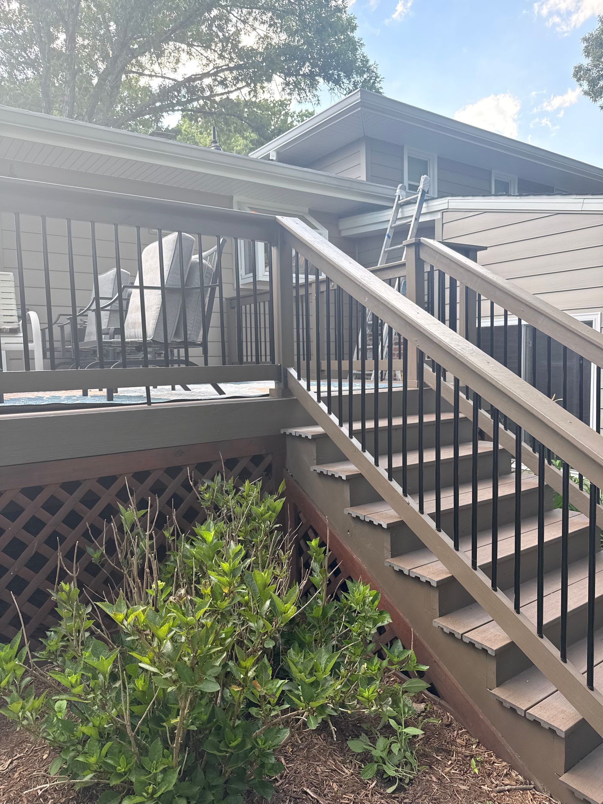 Wooden deck stairs with black railings beside a house, with shrubs and sunlight.