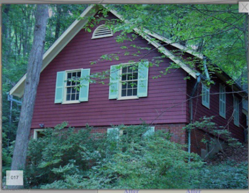 Red wooden house with white shutters, partly hidden by trees and shrubs in a wooded yard