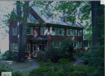Large dark wooden house with white trim, partly hidden by trees and greenery in a shaded yard