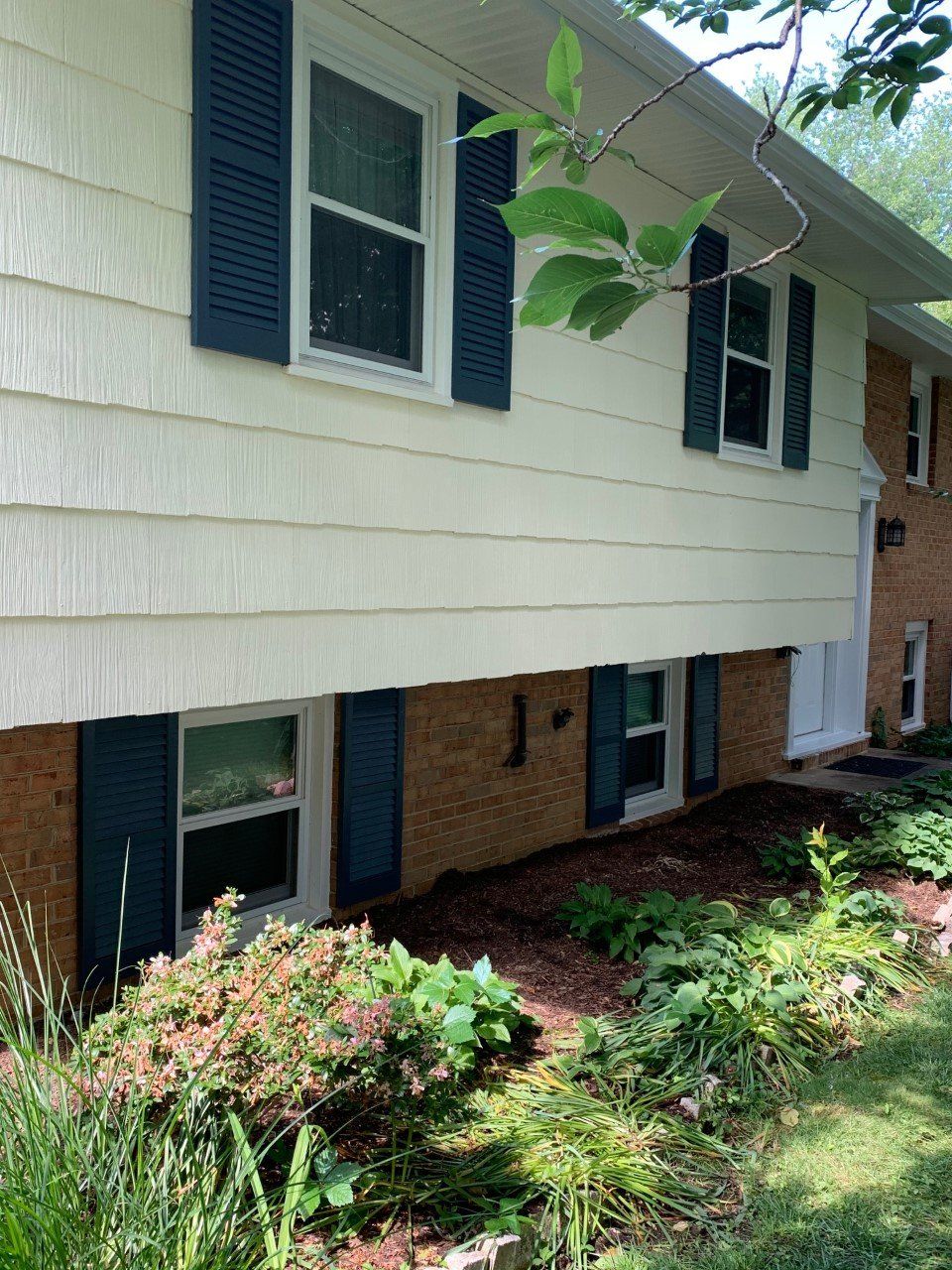 Suburban house exterior with white siding, blue shutters, brick lower level, and a landscaped front garden.