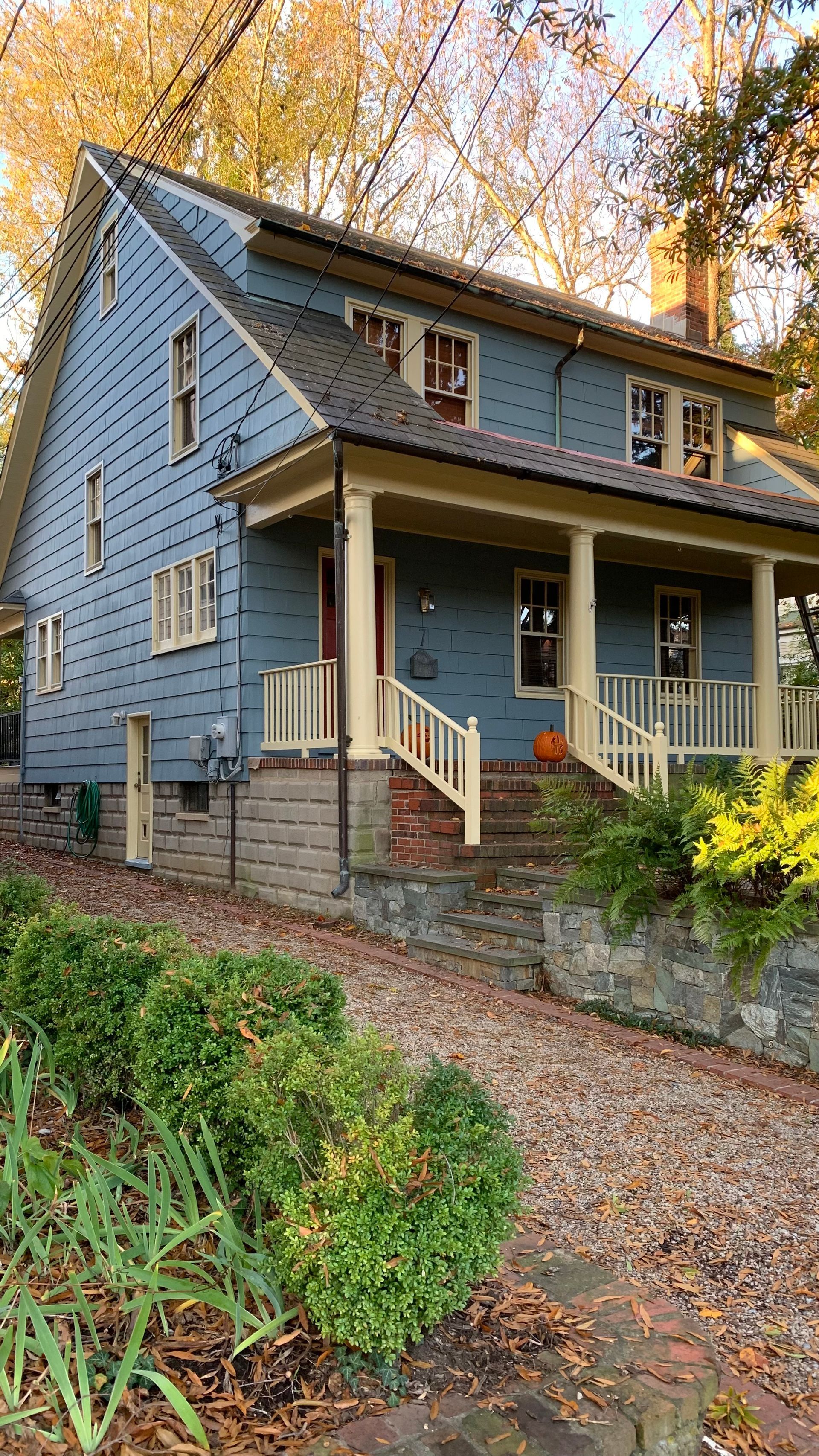 Blue-gray house with a covered porch and autumn foliage beside a gravel driveway