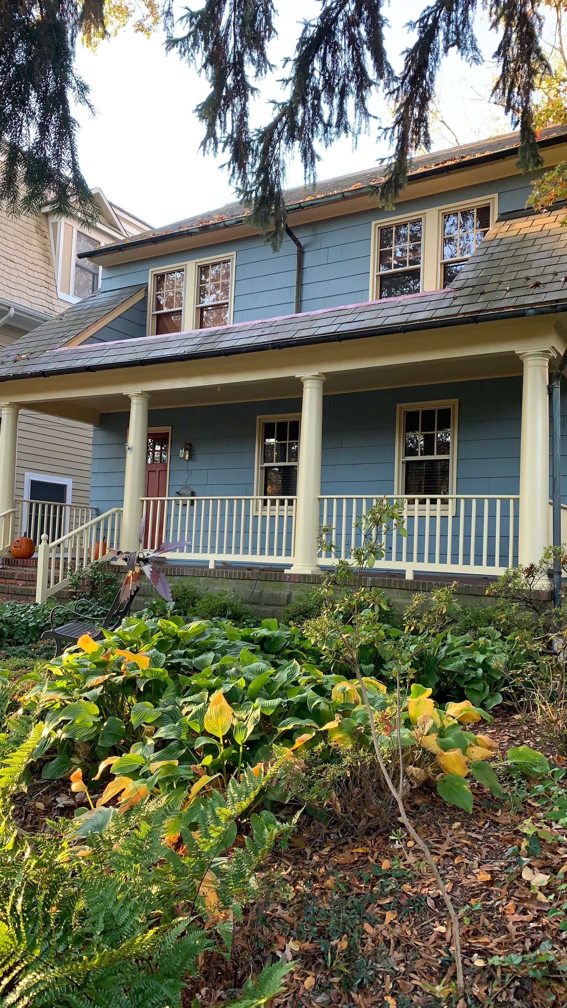 Blue two-story house with white porch columns and railings, surrounded by trees and a flowering garden.