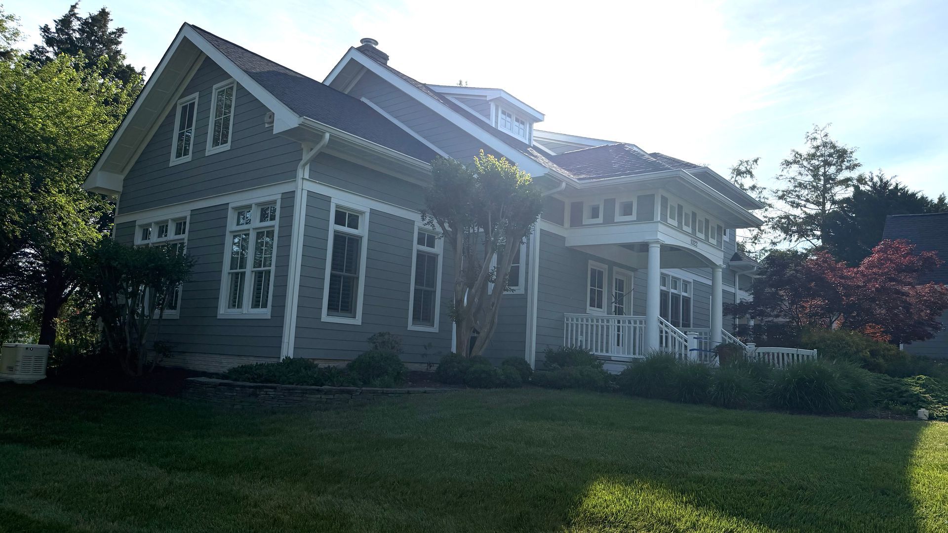 Gray house with white trim and porch in a sunny yard, with trees and lawn in front.