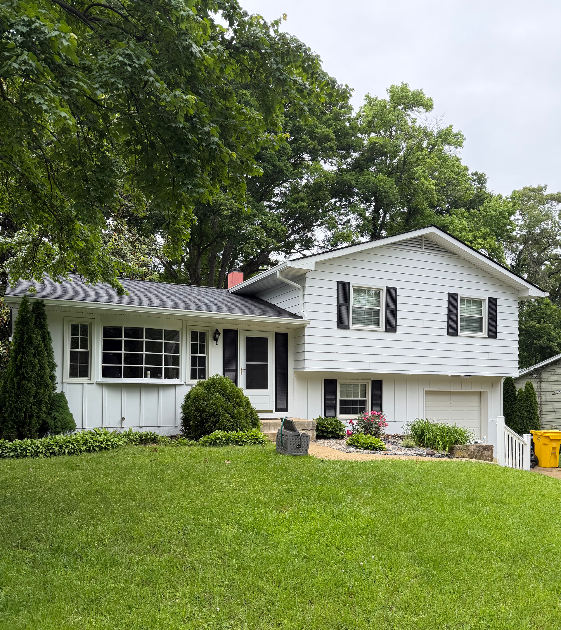 White two-story house with black shutters and a green lawn under large trees