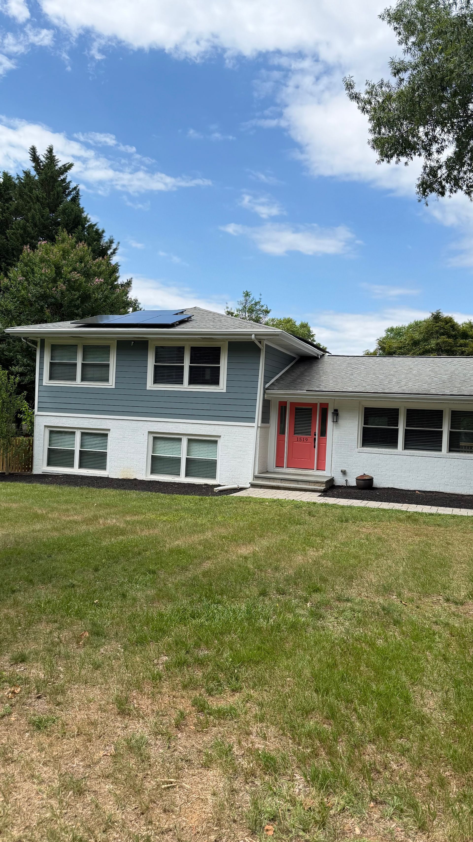 Two-story gray house with white trim, red front door, and a grassy yard under a partly cloudy sky