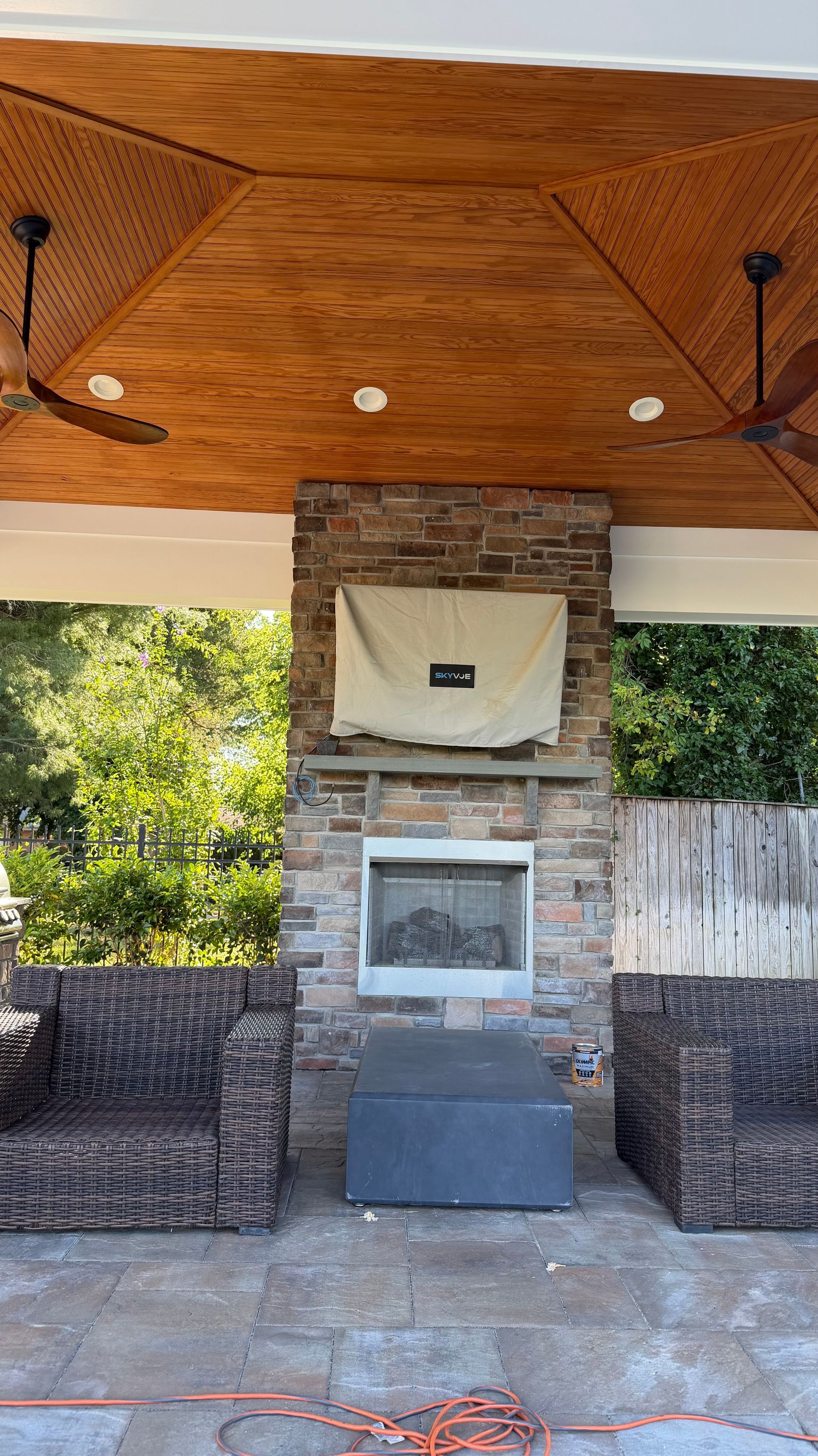 Outdoor stone fireplace with a wood-paneled ceiling and wicker chairs on a patio.