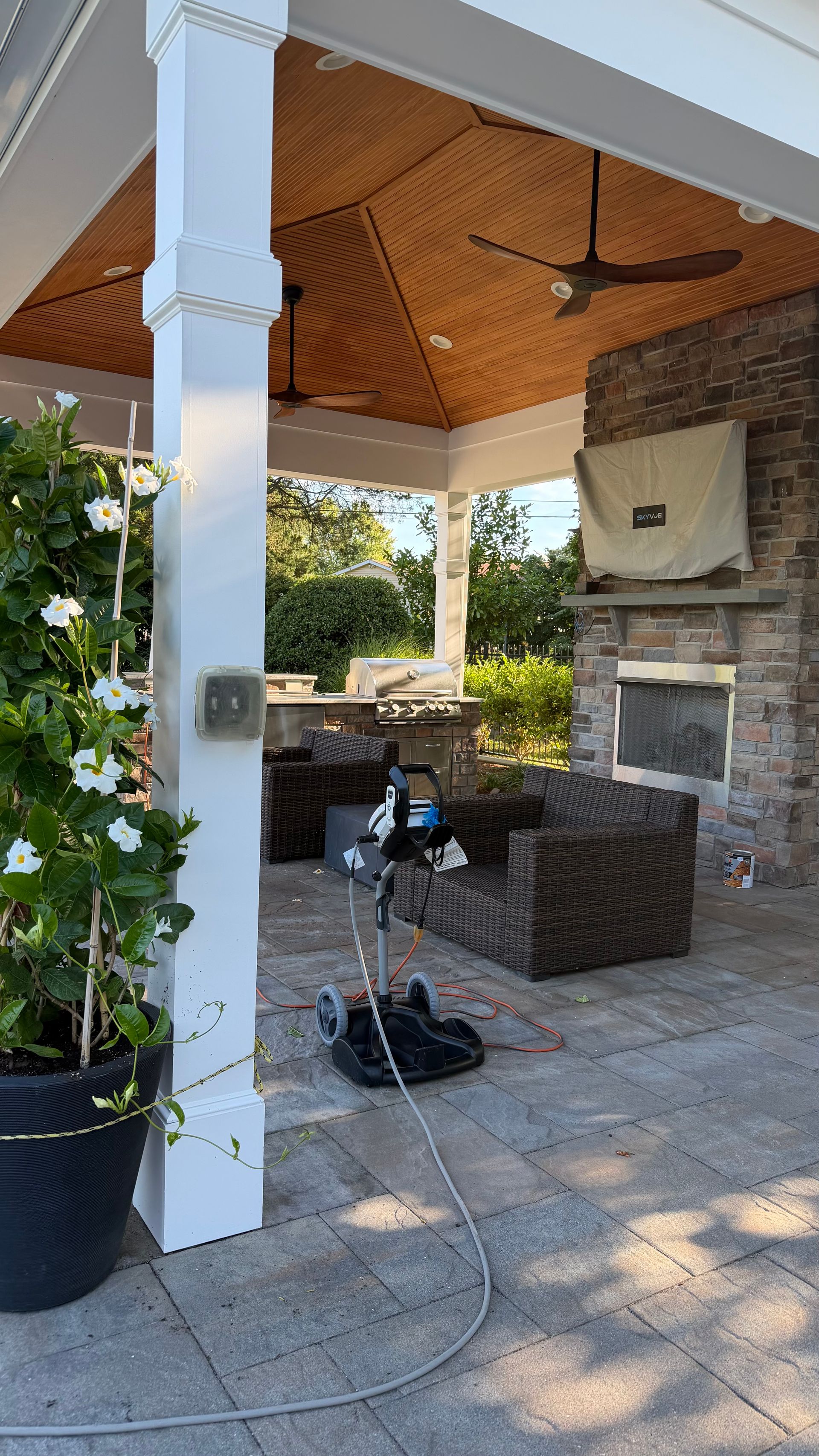 Covered patio with a wicker chair, ceiling fan, stone fireplace, and potted plants.