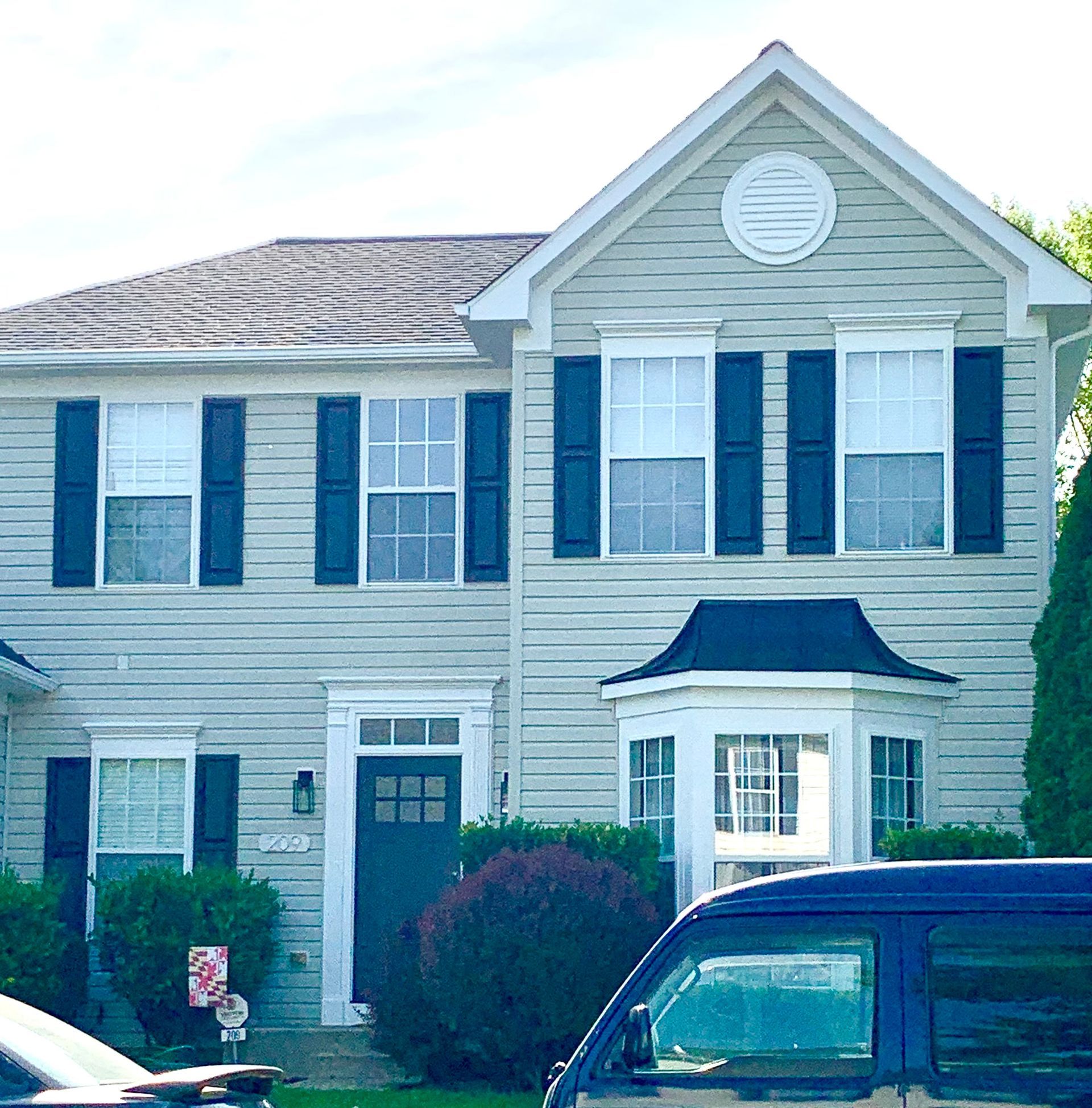 Two-story beige house with blue shutters, white trim, and a parked dark vehicle in front