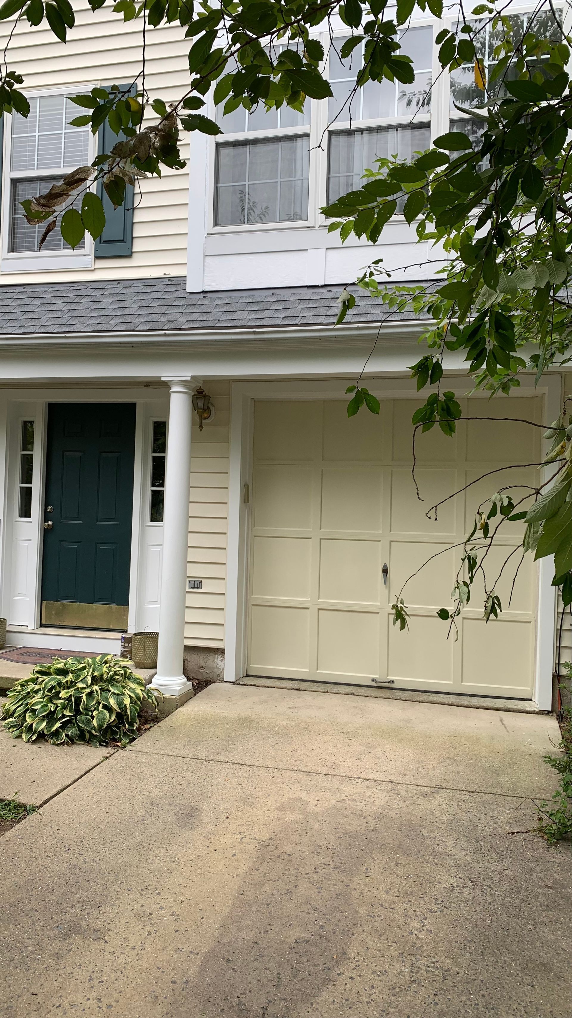 Suburban house exterior with green front door, white siding, garage, and driveway bordered by leafy trees