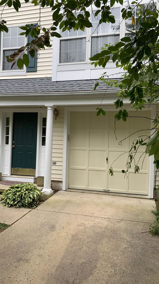 Front of a townhouse with a green door, beige garage, and a short driveway, framed by leafy branches