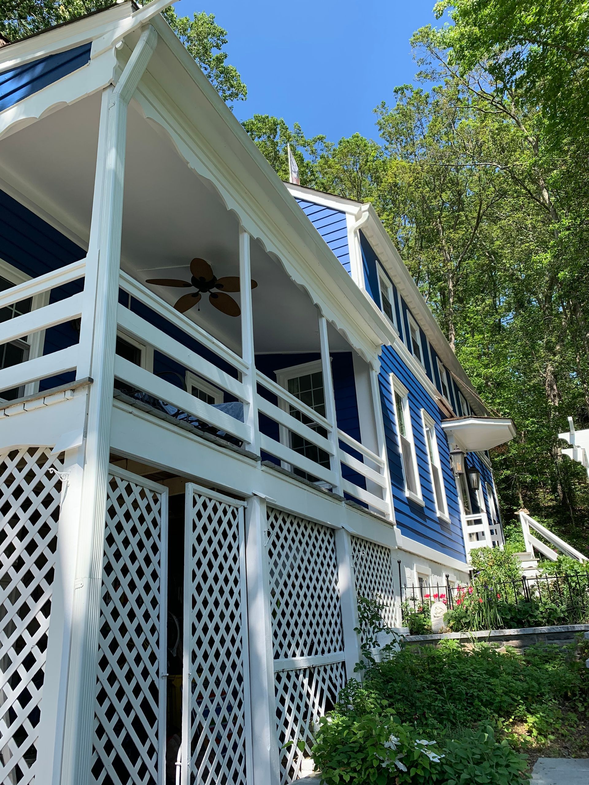 White porch and blue-trimmed house beside a leafy hillside under a bright blue sky