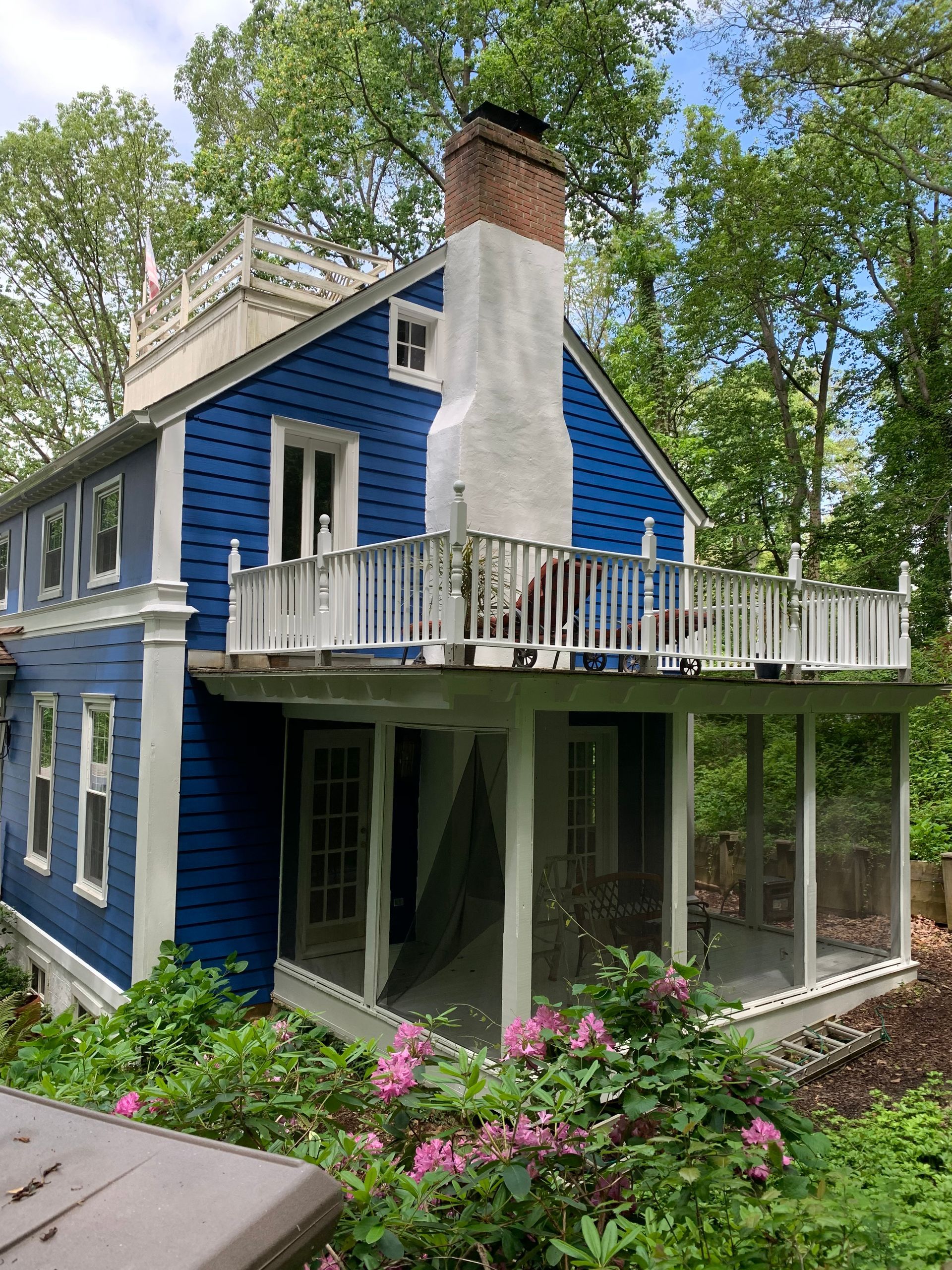 Blue two-story house with white trim, chimney, and wraparound porch surrounded by greenery and pink flowers