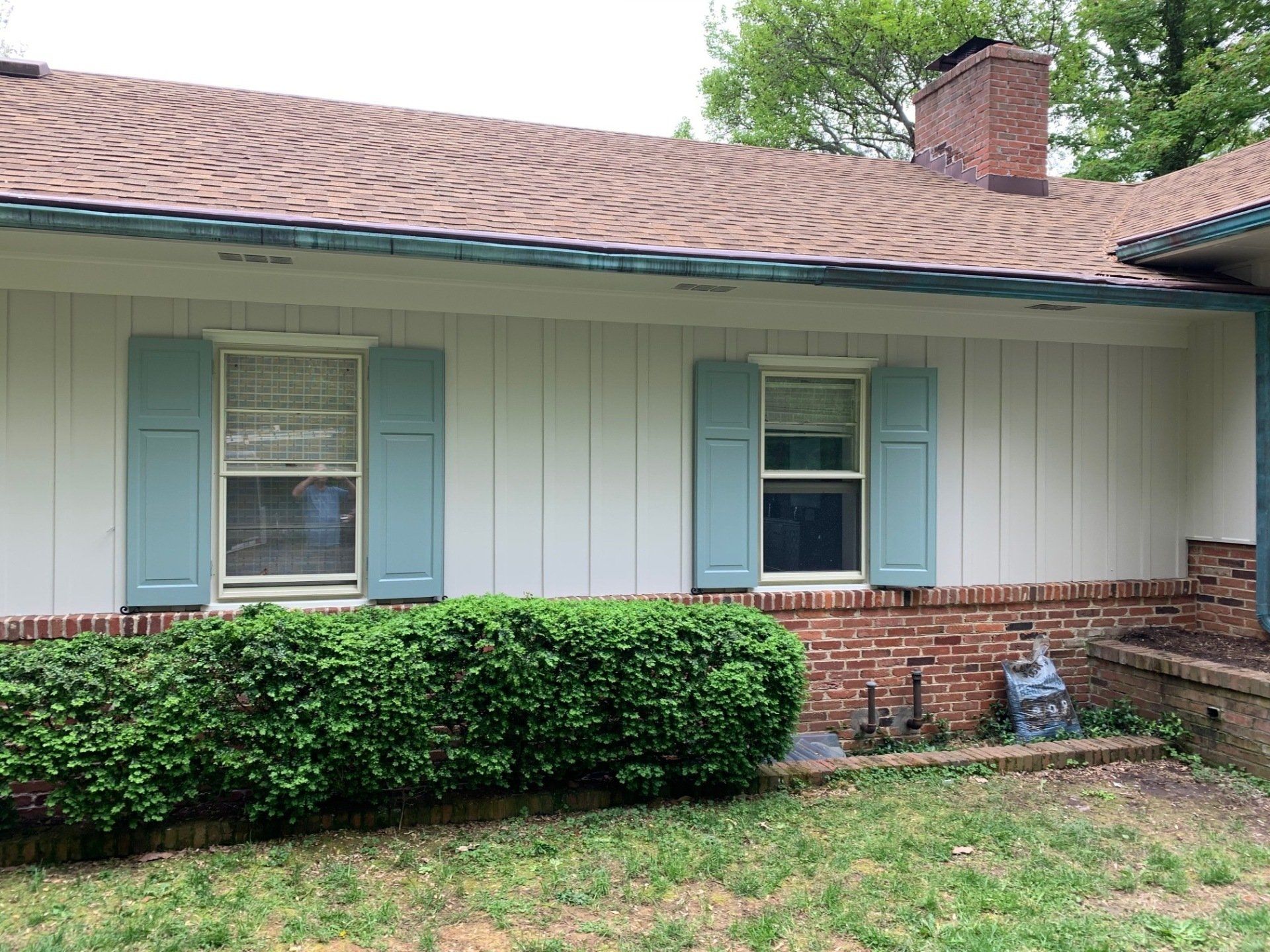 Single-story house exterior with white siding, blue shutters, brick base, and green bushes in front.