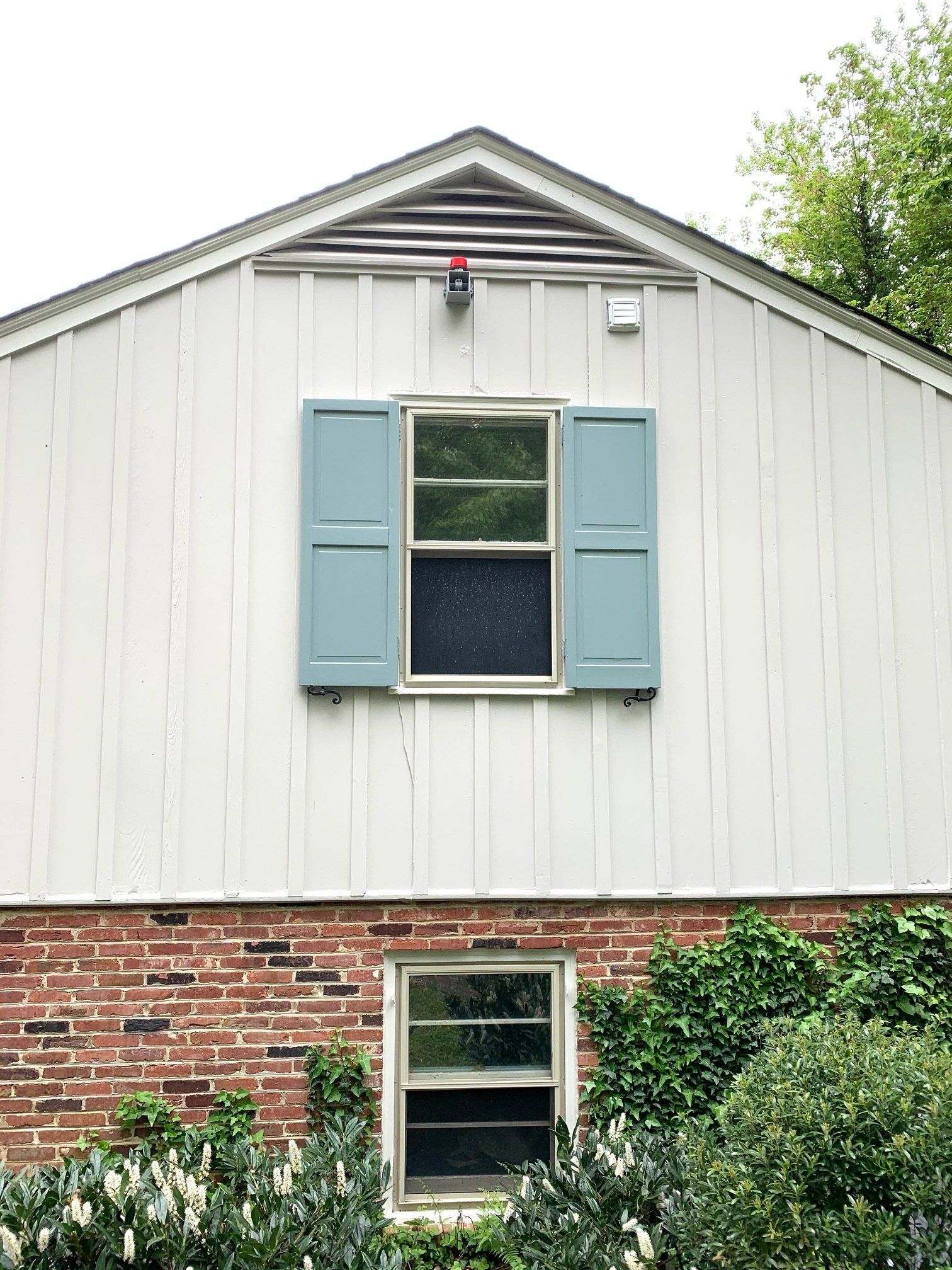 White house exterior with blue shutters, brick lower wall, and two windows with greenery below