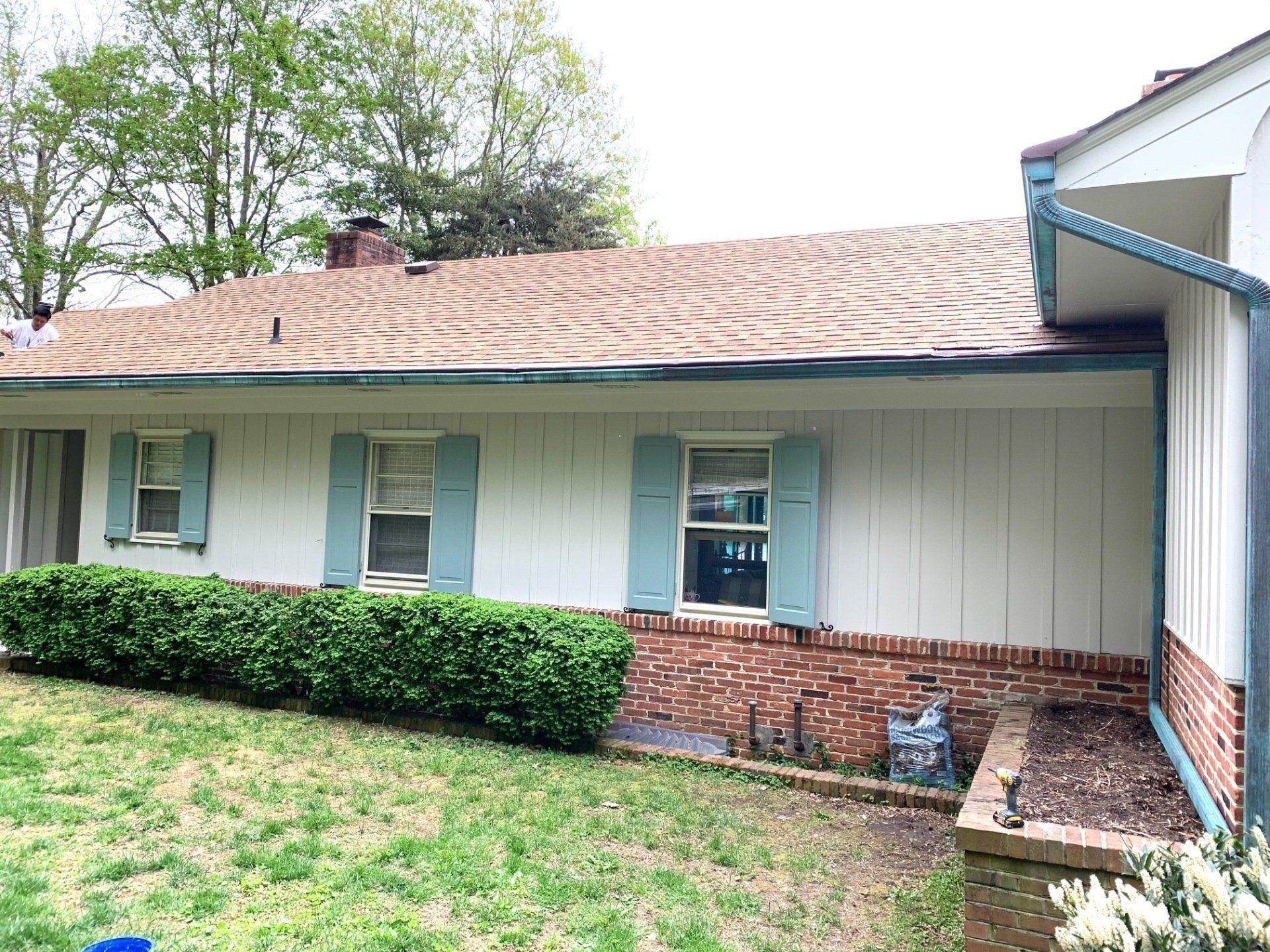 Single-story house exterior with beige siding, blue shutters, brick trim, and a hedge in front