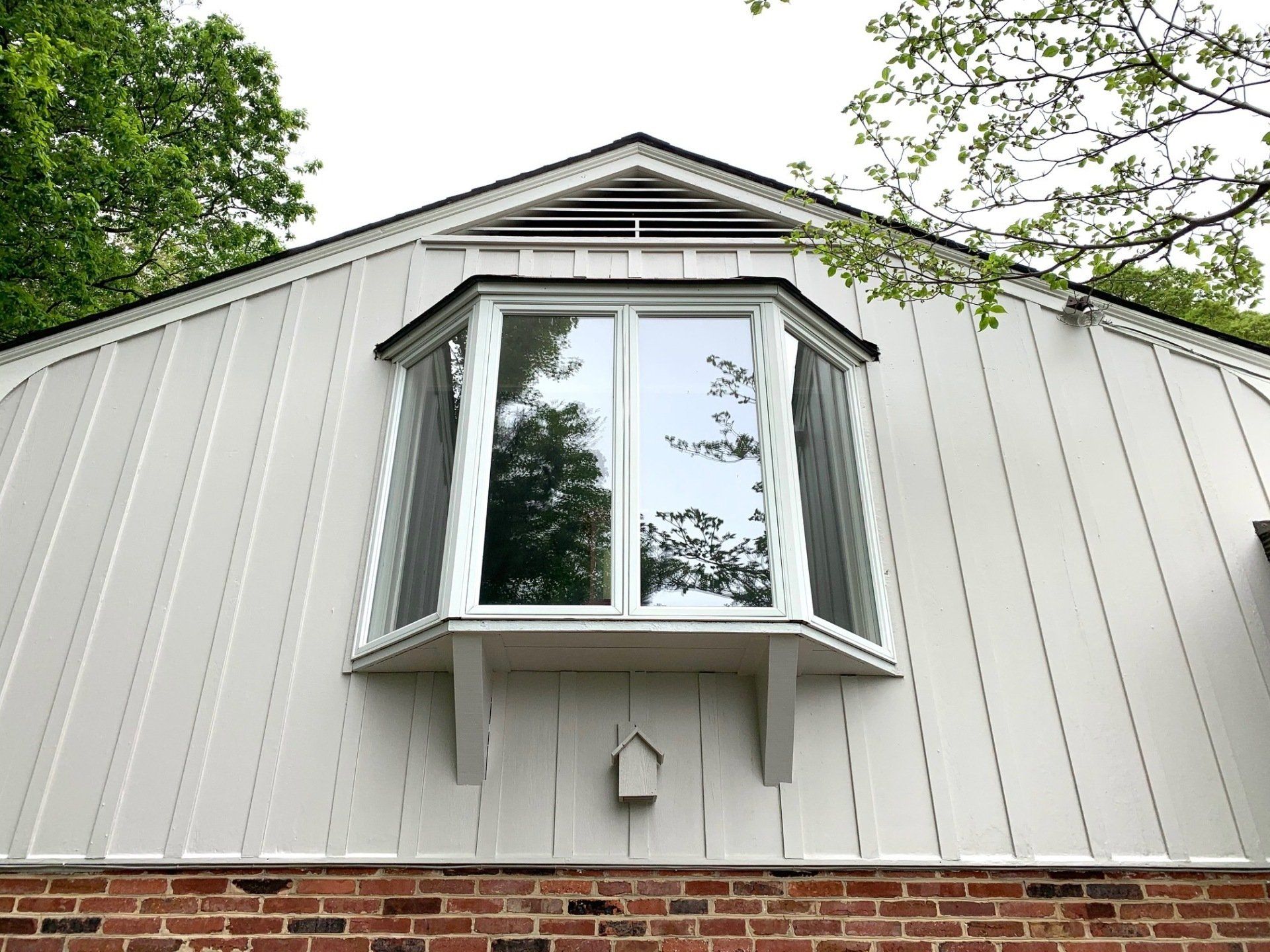 White house exterior with a bay window, green trees reflecting in the glass, and a brick foundation